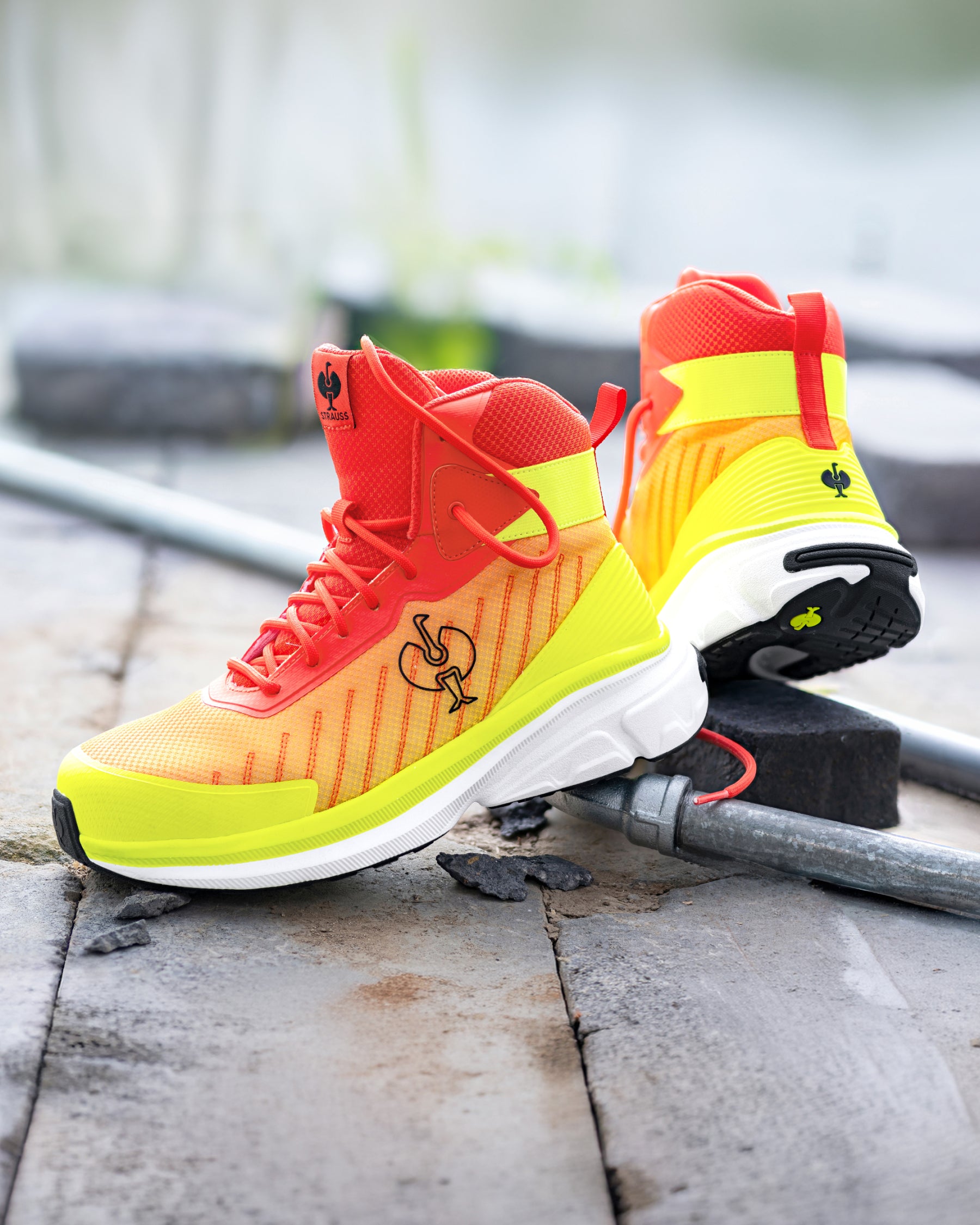 A pair of athletic sneakers in a bright gradient of red, orange, and neon yellow, resting on a weathered stone surface with a metal pipe in the background.