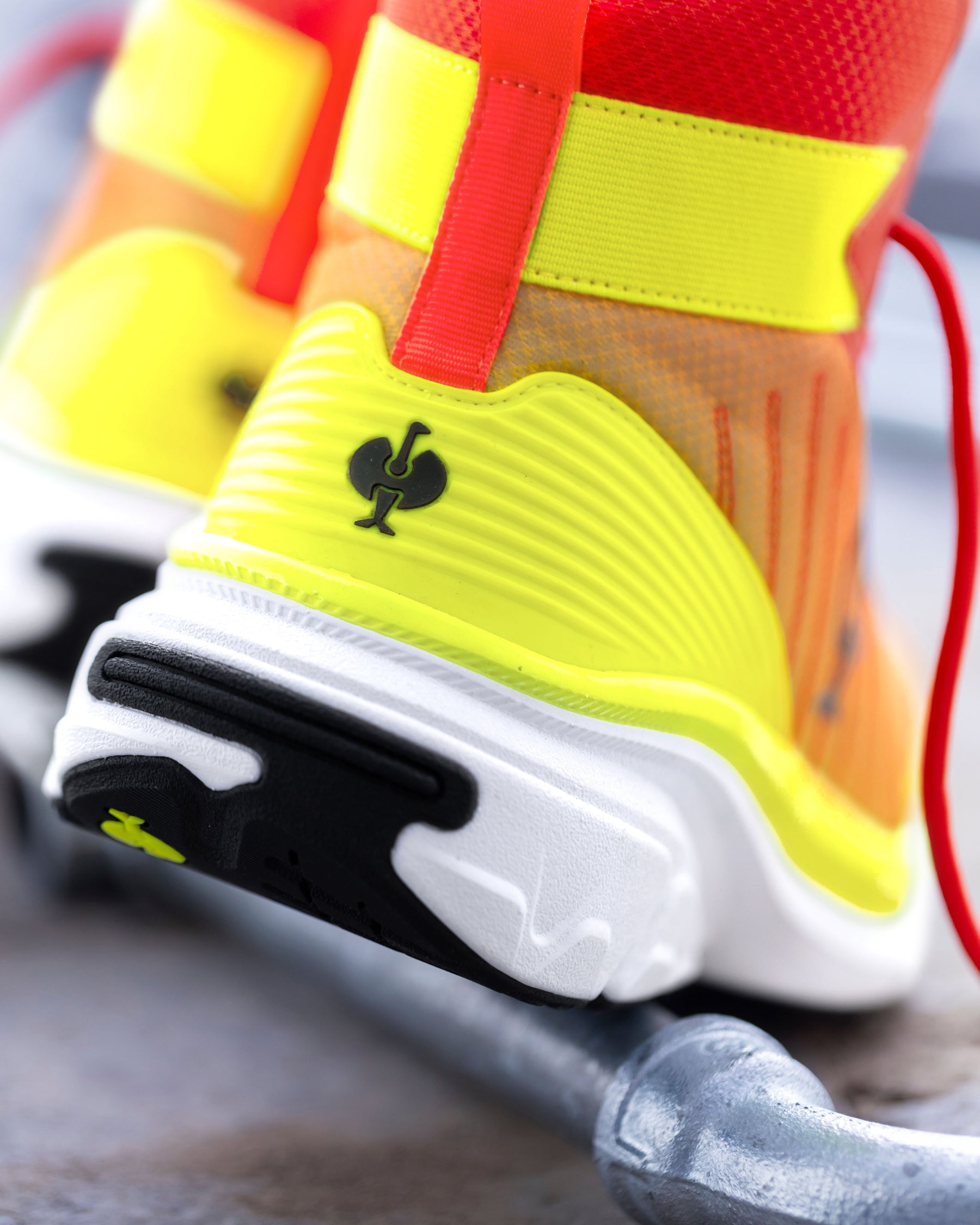 Close-up of a bright neon yellow and orange athletic shoe with red straps, a black logo on the heel, and a white and black sole, resting on a gray metal surface.