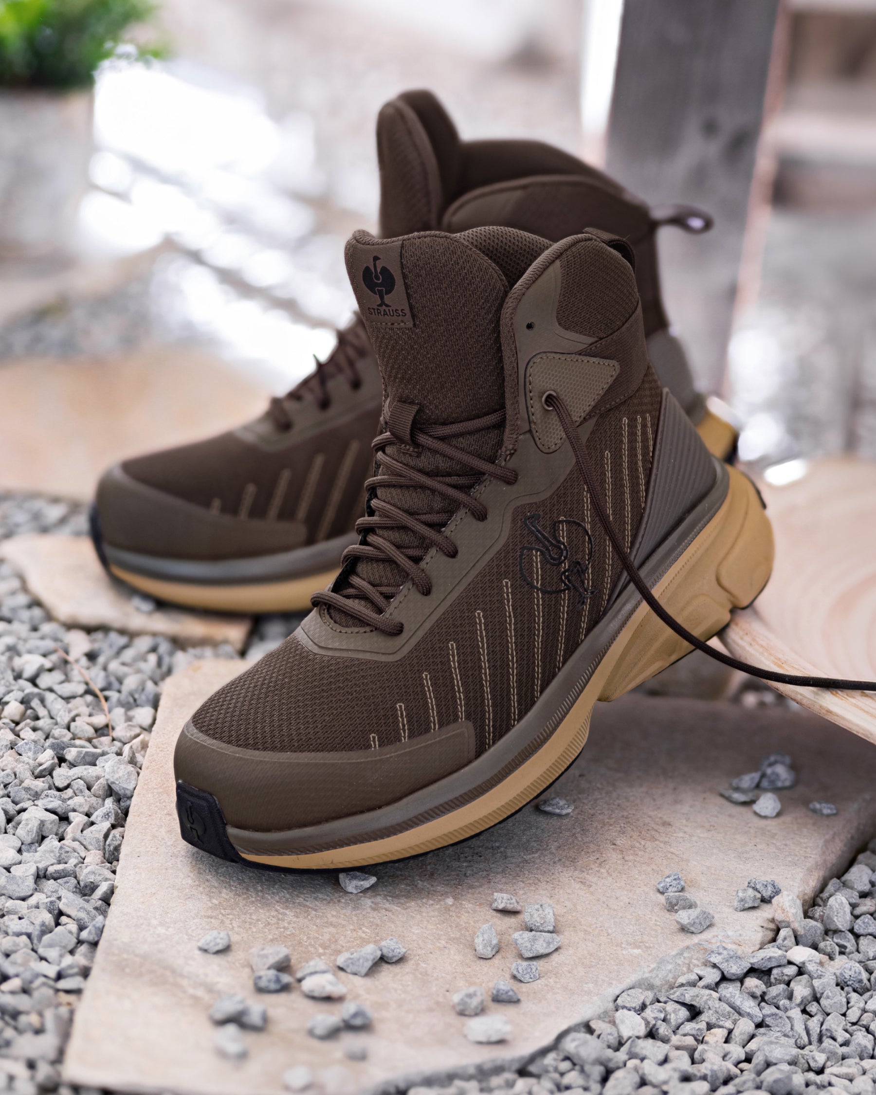 A pair of mid-cut lace-up boots in brown and olive tones with tan soles, photographed outdoors on stone slabs over gravel.