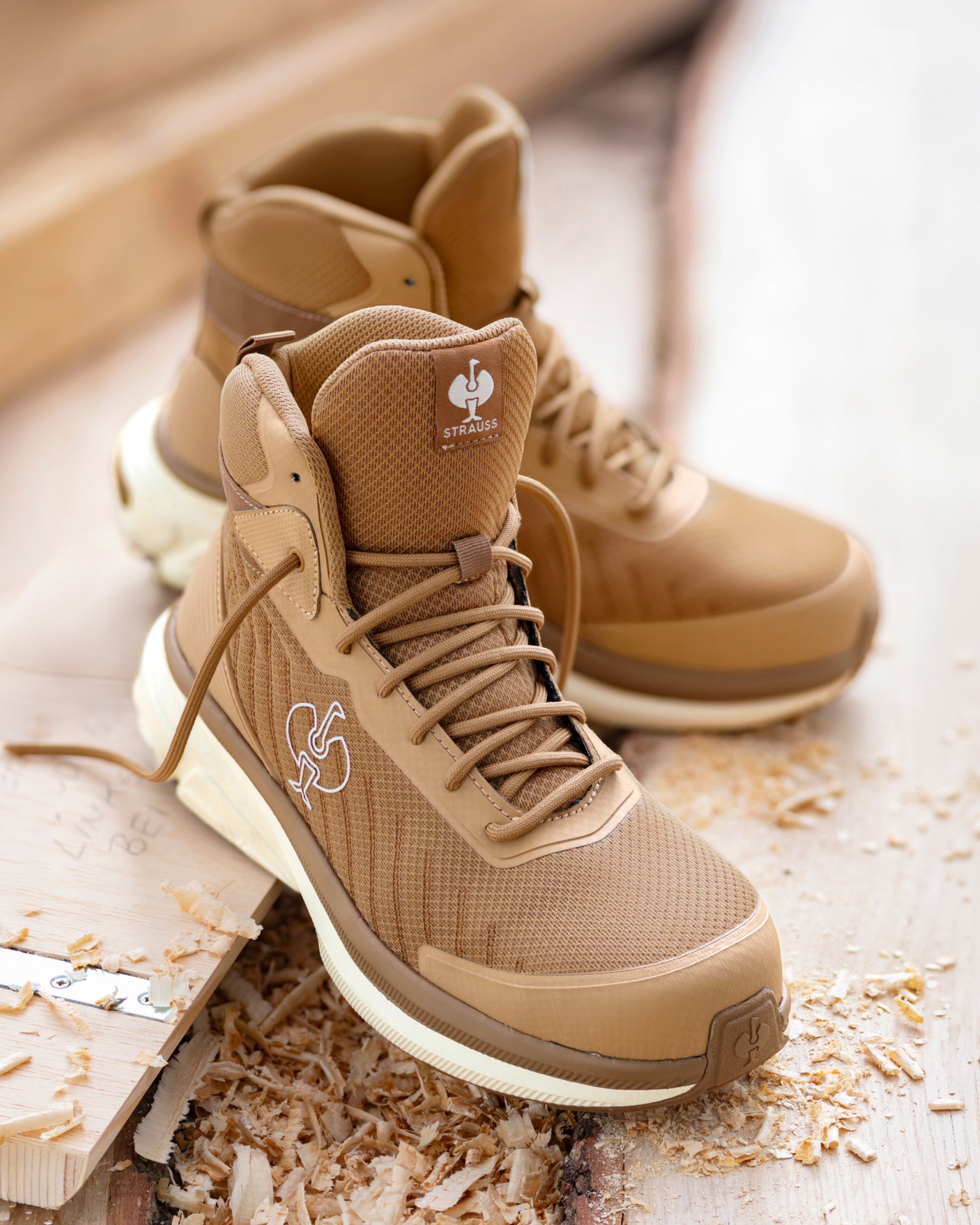 A pair of tan high-top work boots with white midsoles, styled on a wooden surface with wood shavings. The boots feature a textured mesh upper, lace-up front, and a small logo patch on the tongue.