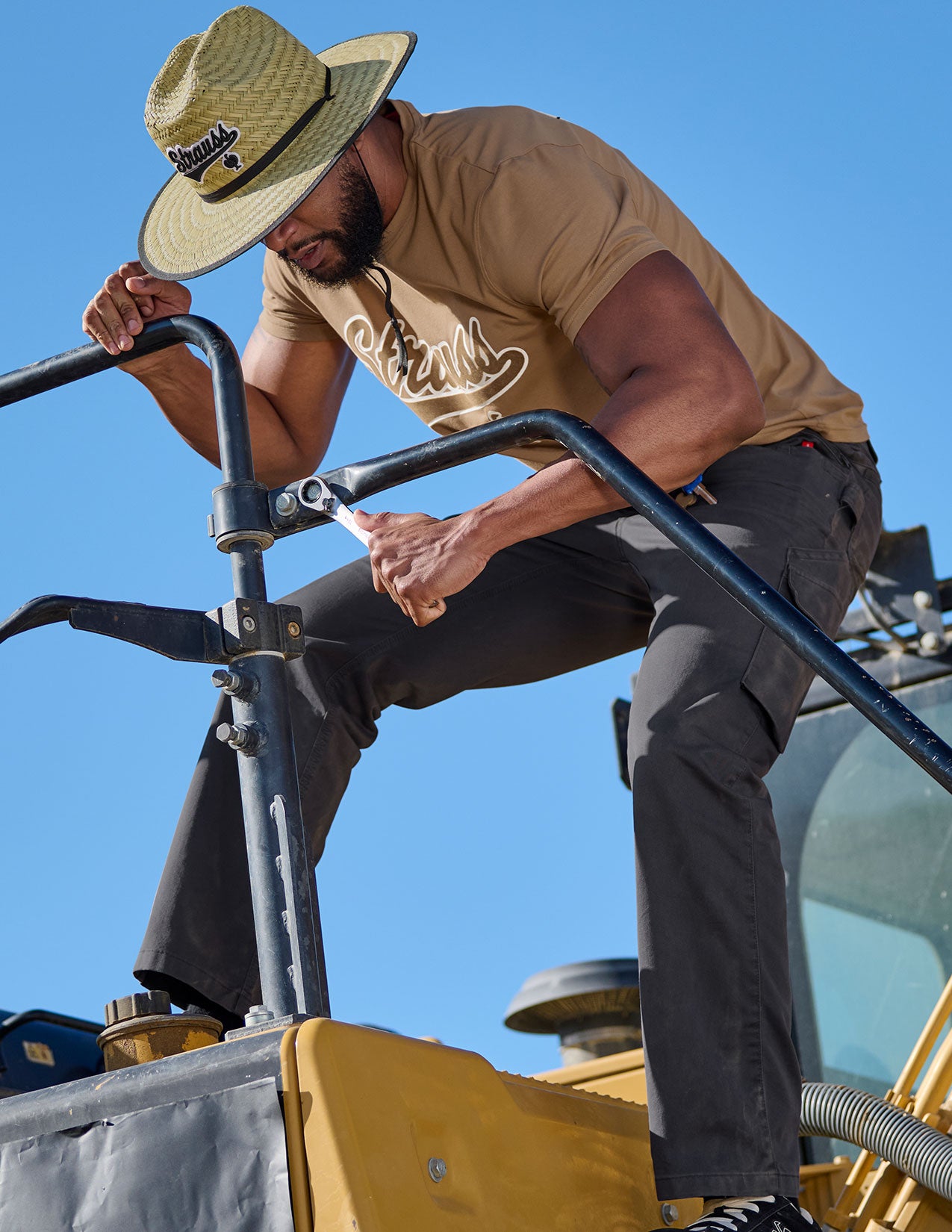 Man standing on a piece of construction equipment against a clear blue sky and wearing a straw hat.