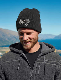 Man wearing a black beanie with 'Strauss' logo outdoors against a blue sky and mountains.