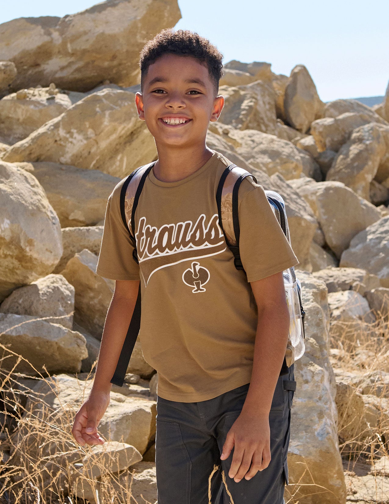 Boy with a clear backpack standing among rocks in a natural setting.