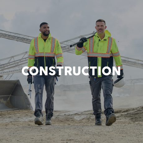 Two construction workers in high-visibility jackets walking on a construction site with the word 'CONSTRUCTION' displayed.