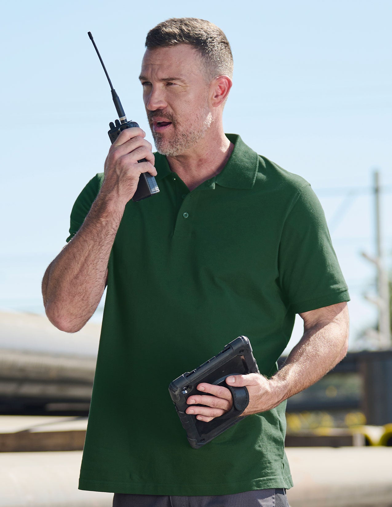 Man in a green polo shirt holding a walkie-talkie and tablet outdoors.