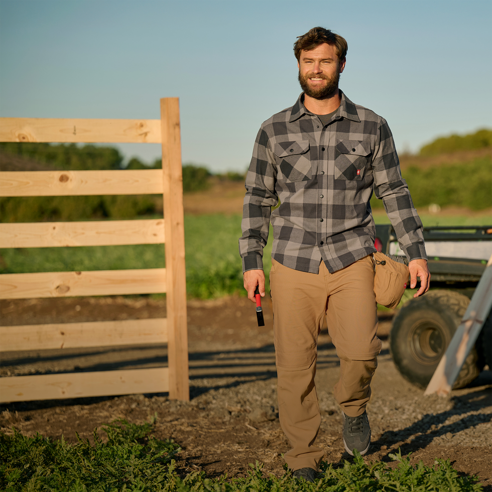 Man standing on a farm holding a tool, wearing a plaid shirt and tan pants.