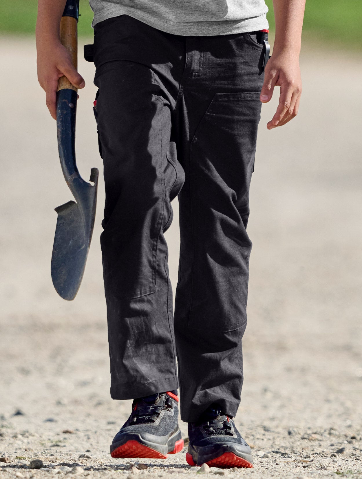 Child wearing black pants and red shoes holding a shovel on a gravel path.