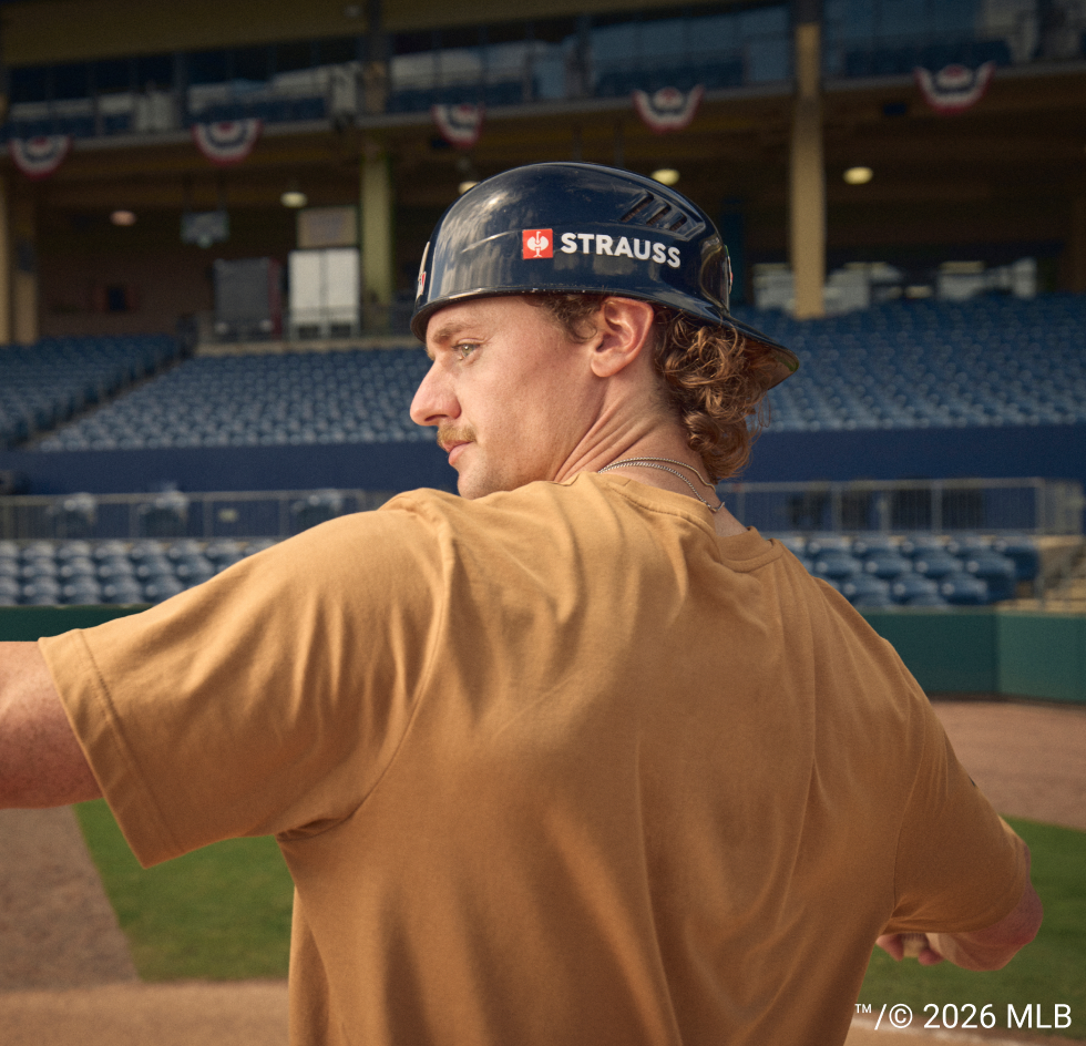 A man wearing a black Strauss batting helmet and a tan t-shirt, photographed from behind in a stadium with blue seats in the background.