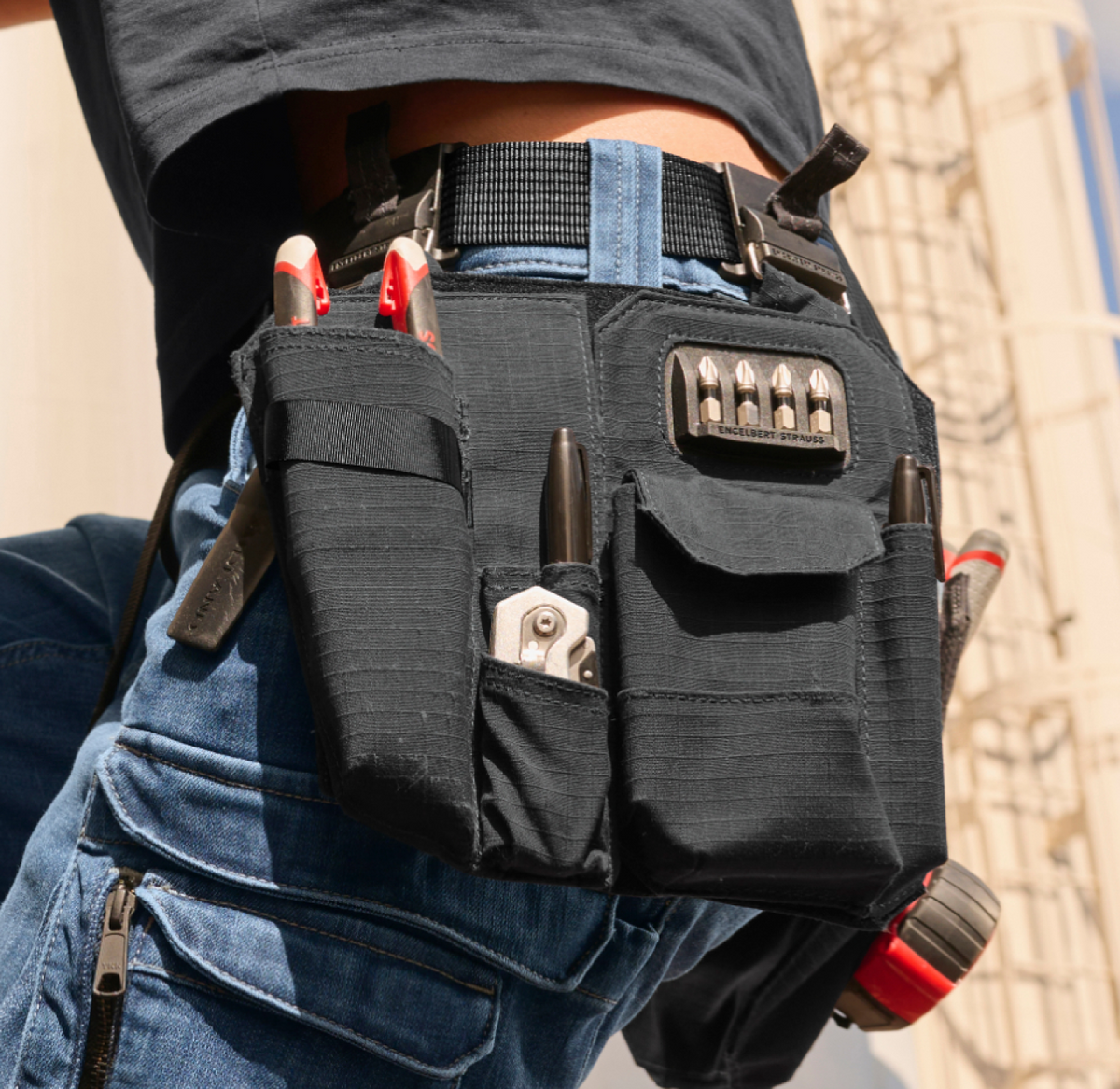 A close-up of a person wearing blue jeans and a black tool belt filled with various tools, including pliers, screwdrivers, and a tape measure, against an industrial outdoor background.