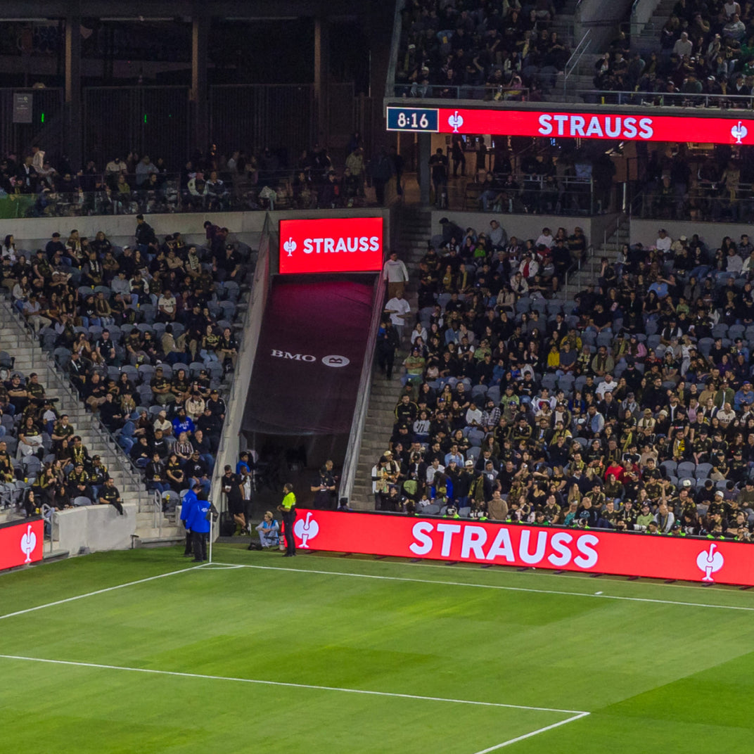 A wide view of a crowded soccer stadium with a green field, stands filled with spectators, and large red STRAUSS advertising boards around the pitch.