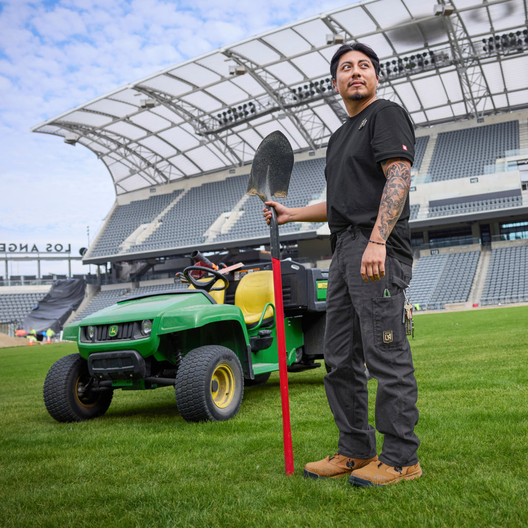 A man stands on a green sports field holding a shovel in front of a stadium, with a green John Deere utility vehicle behind him.