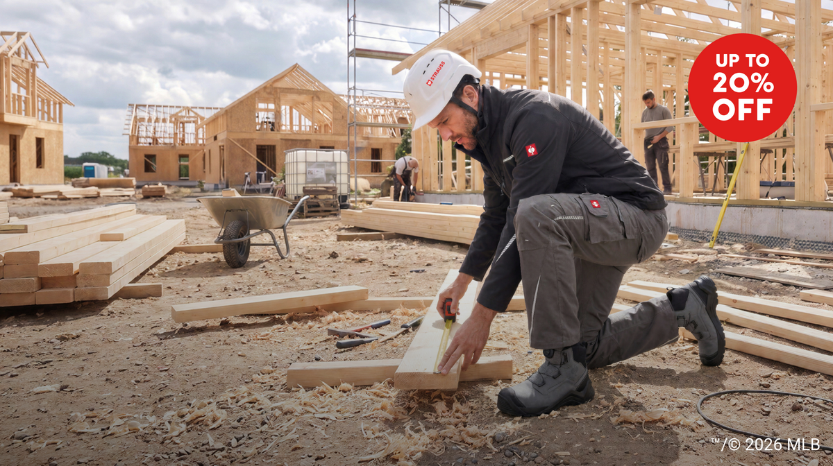 A man in STRAUSS workwear, including a white hard hat, a black softshell jacket, and gray work pants, measures wood on a construction site. Other workers and wooden houses are visible in the background under a cloudy sky, with a red 'UP TO 20% OFF' promotional badge in the top right.