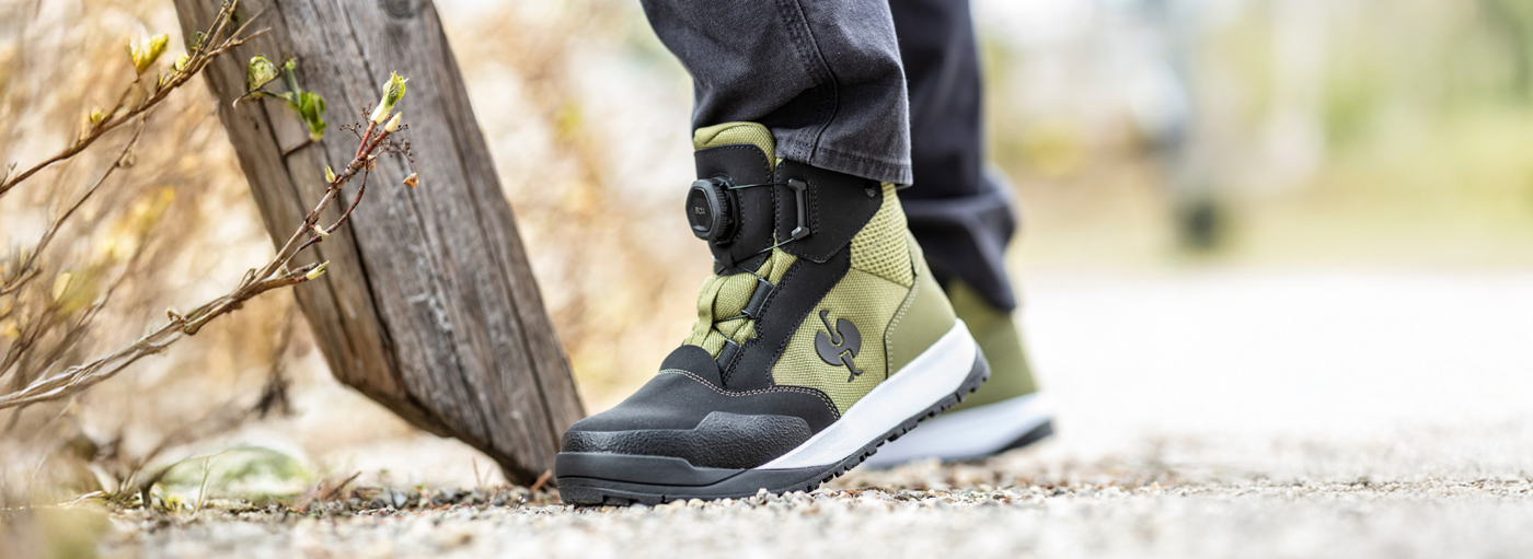 A close-up of a person's foot in stylish green and black boots, standing on a gravel path.