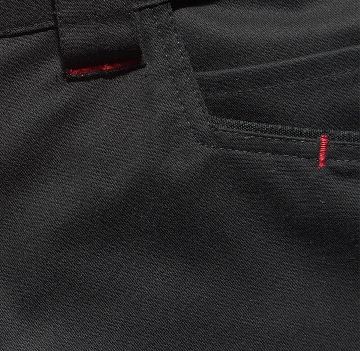 Close-up of black fabric pants with visible stitching and pocket. There are small red thread details on the belt loop and pocket corner, contrasting with the dark material.
