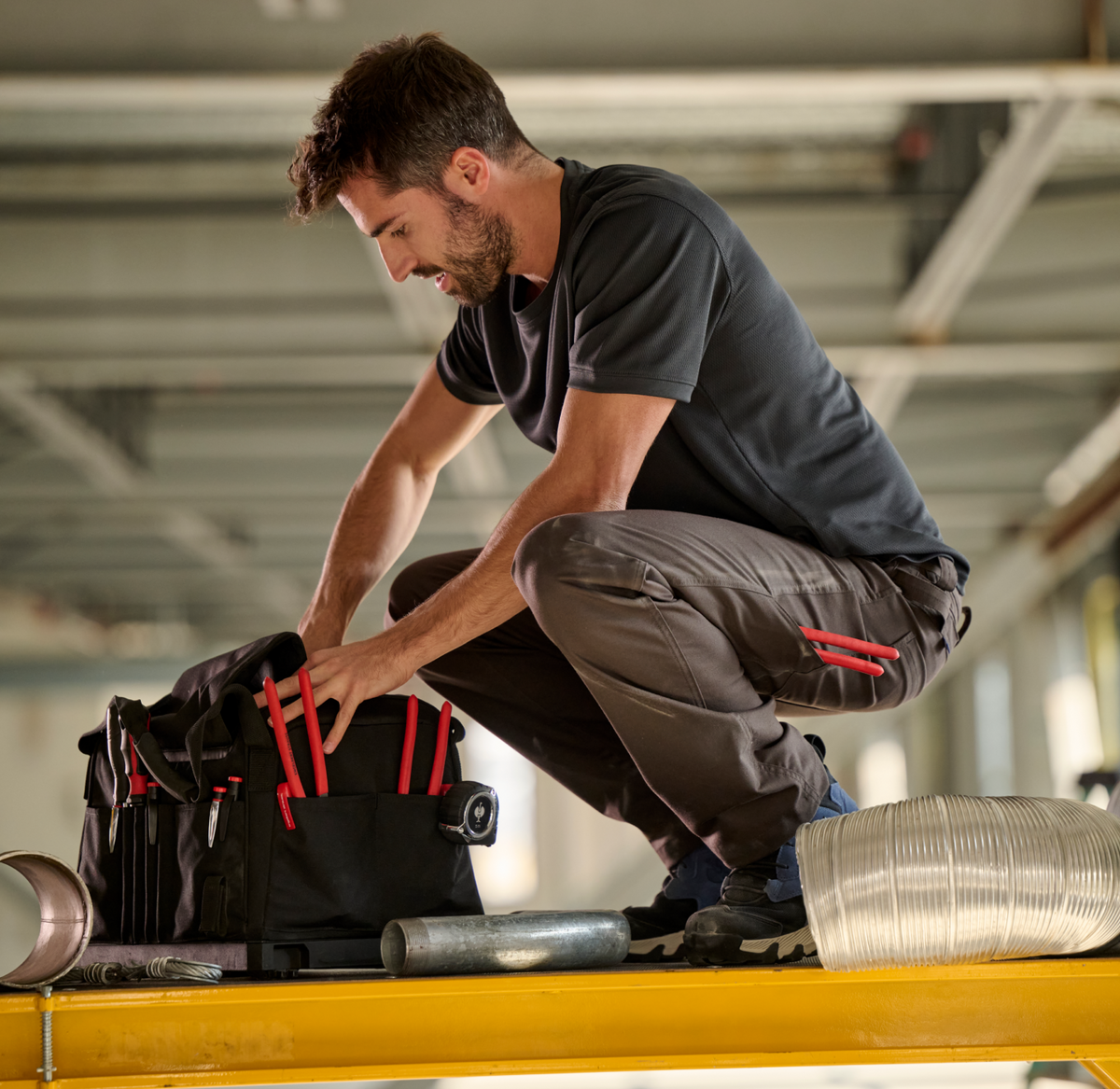 A person in work clothes crouches on a yellow platform, reaching into a black tool bag filled with red-handled tools, with metal pipes and a flexible duct nearby in an industrial setting.