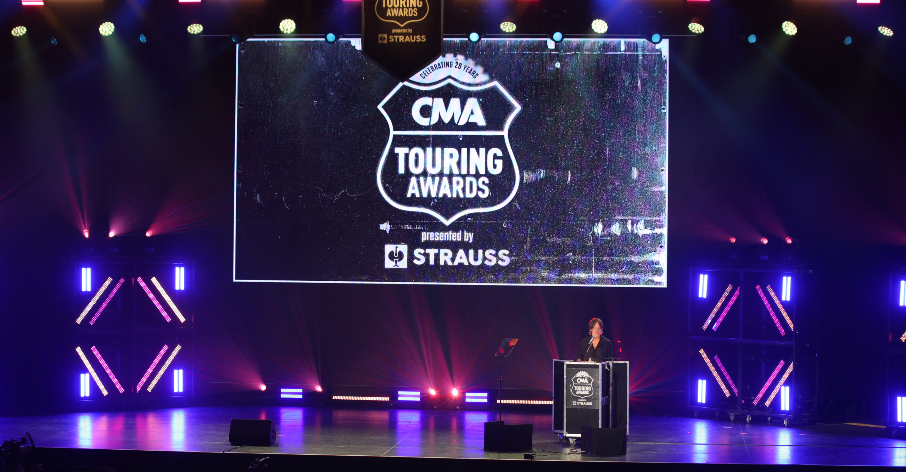 Kieth Urban stands at a podium on the stage of the CMA Touring Awards, presented by STRAUSS