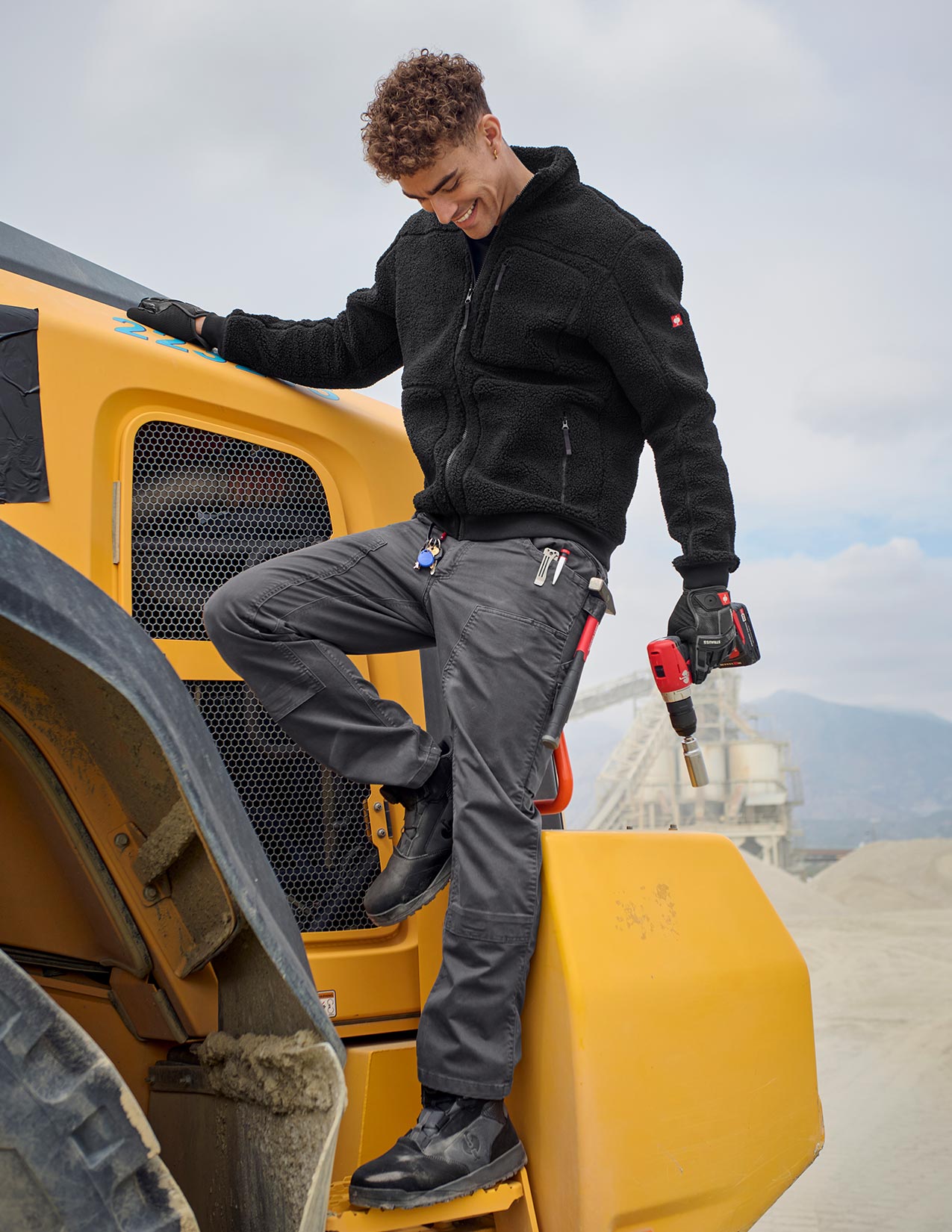 Person in black jacket and gray pants standing on a large yellow piece of machinery with a mountainous background.