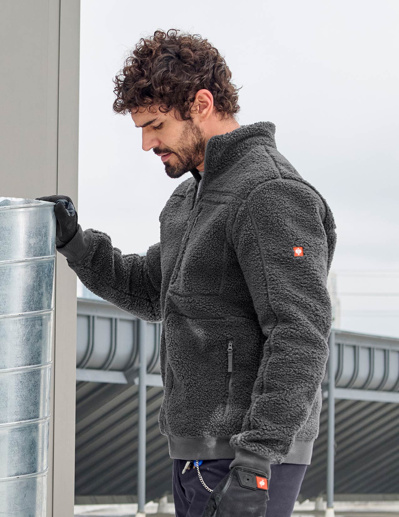 Man wearing a gray fleece jacket with a brand logo, standing next to a metal cylindrical object.