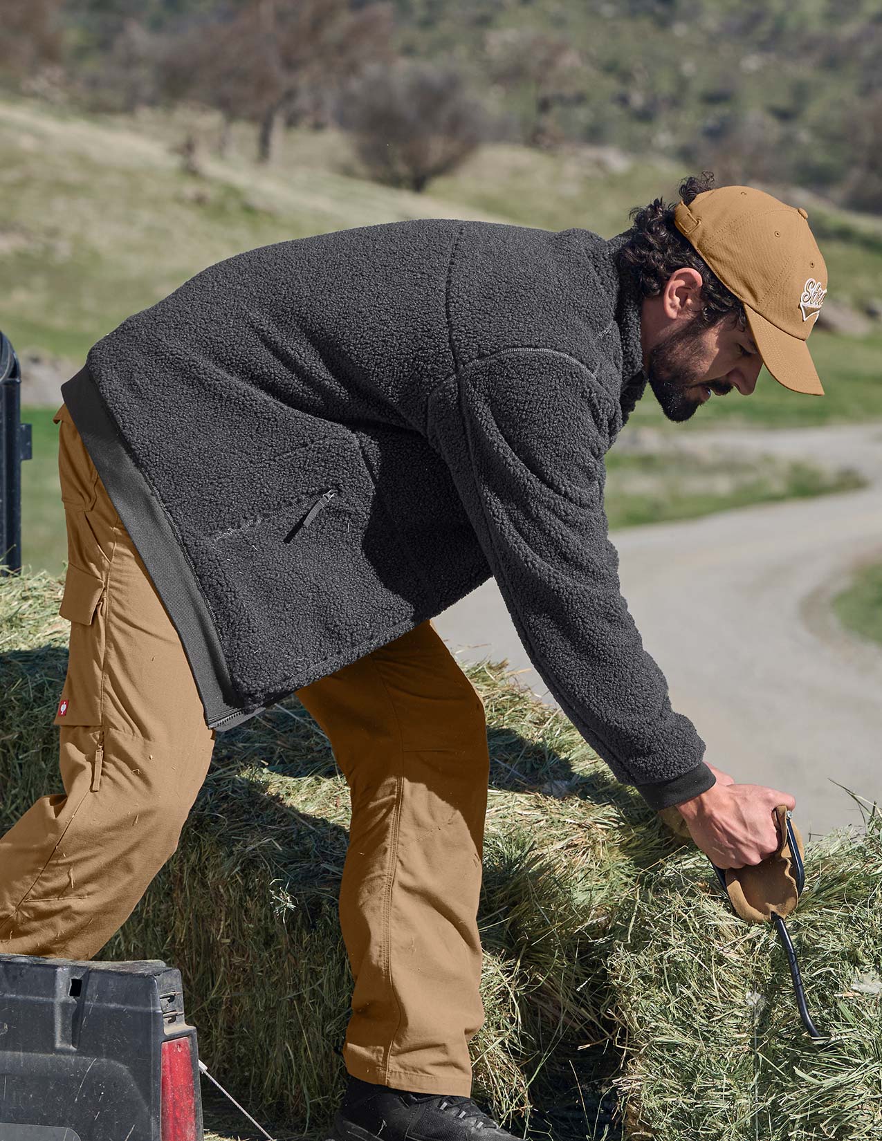 Person in outdoor setting, bending over a stack of hay wearing a gray jacket and tan pants.