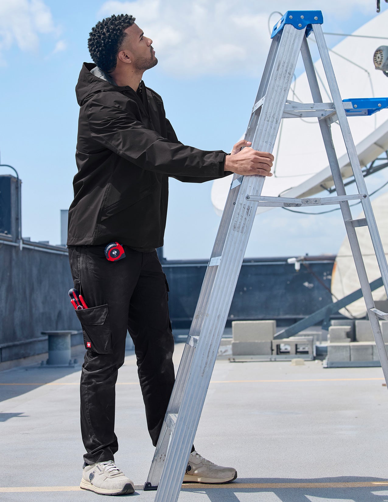 Person on a rooftop holding a ladder, wearing a black jacket and pants with a tool belt.