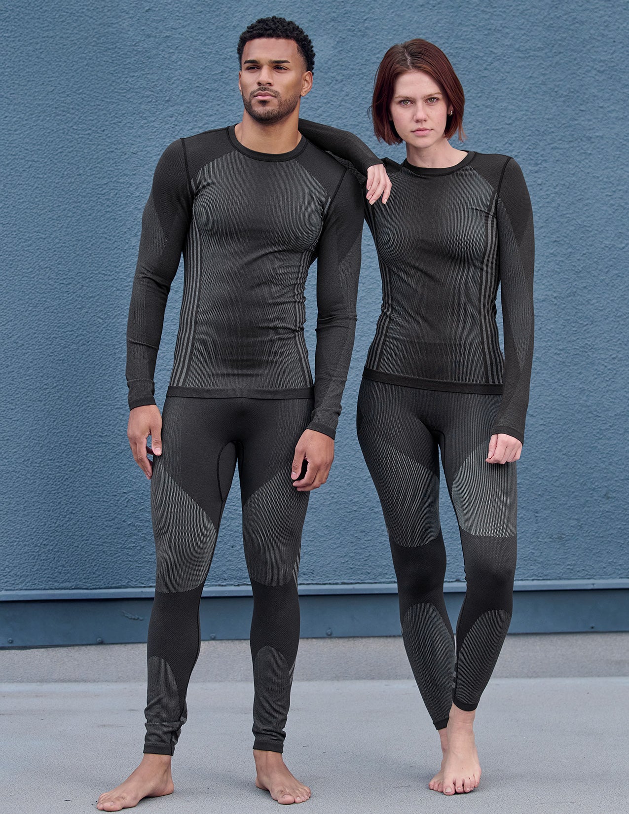 Two people wearing black and gray thermal pants and longsleeves standing against a blue wall.