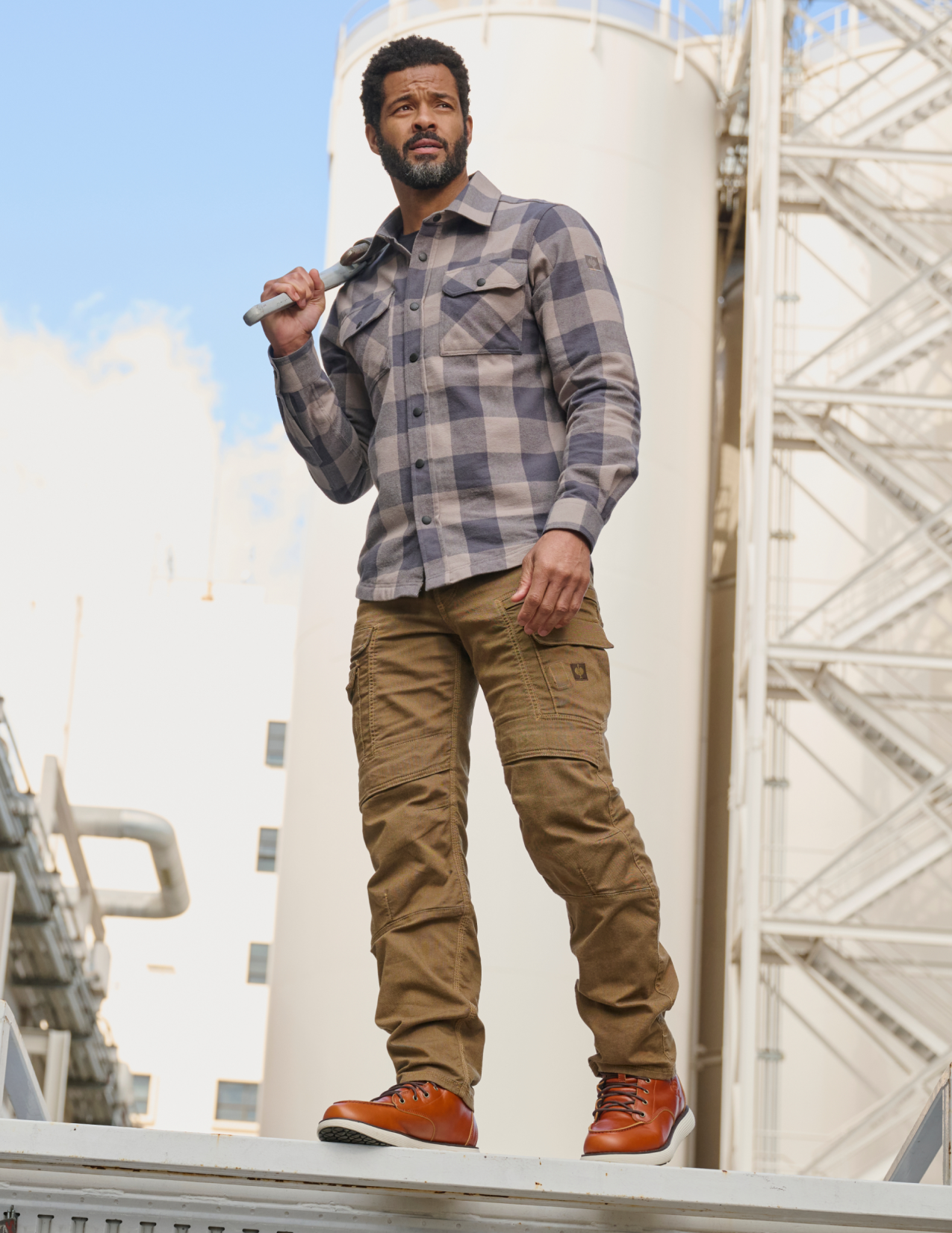 Man wearing a plaid shirt and brown pants standing in front of industrial structures.