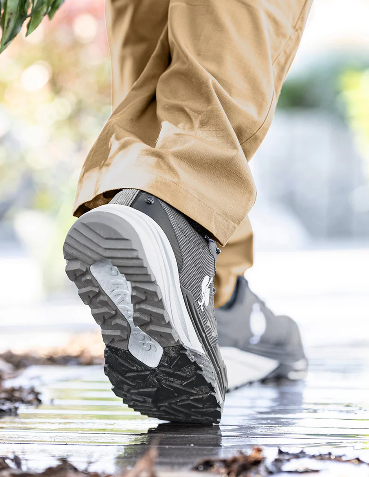 Person wearing brown pants and gray shoes with gray and black sole walking on a wet surface.