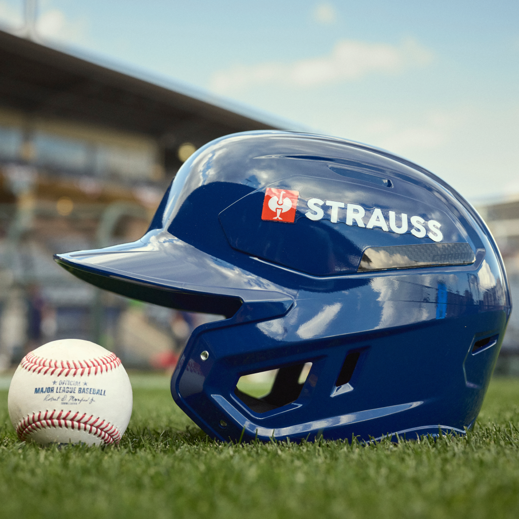 A blue Strauss baseball helmet resting on green grass next to a baseball on a field, with a blurred stadium background.
