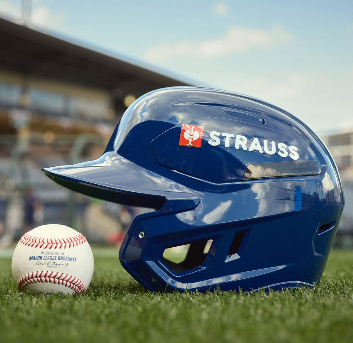 A glossy blue baseball helmet with the STRAUSS branding on the side of it sits on green grass next to a white baseball with red stitching, with a blurred stadium background under a blue sky.