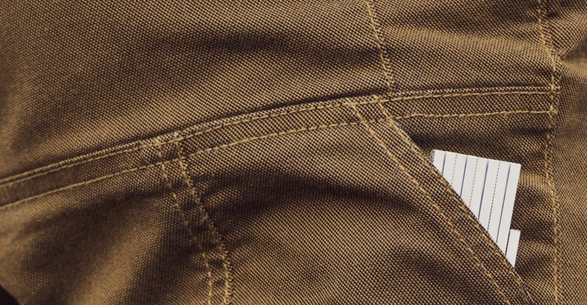 Close-up of brown pants pocket with a piece of white, lined paper partially sticking out. The denim fabric texture and stitching are clearly visible.