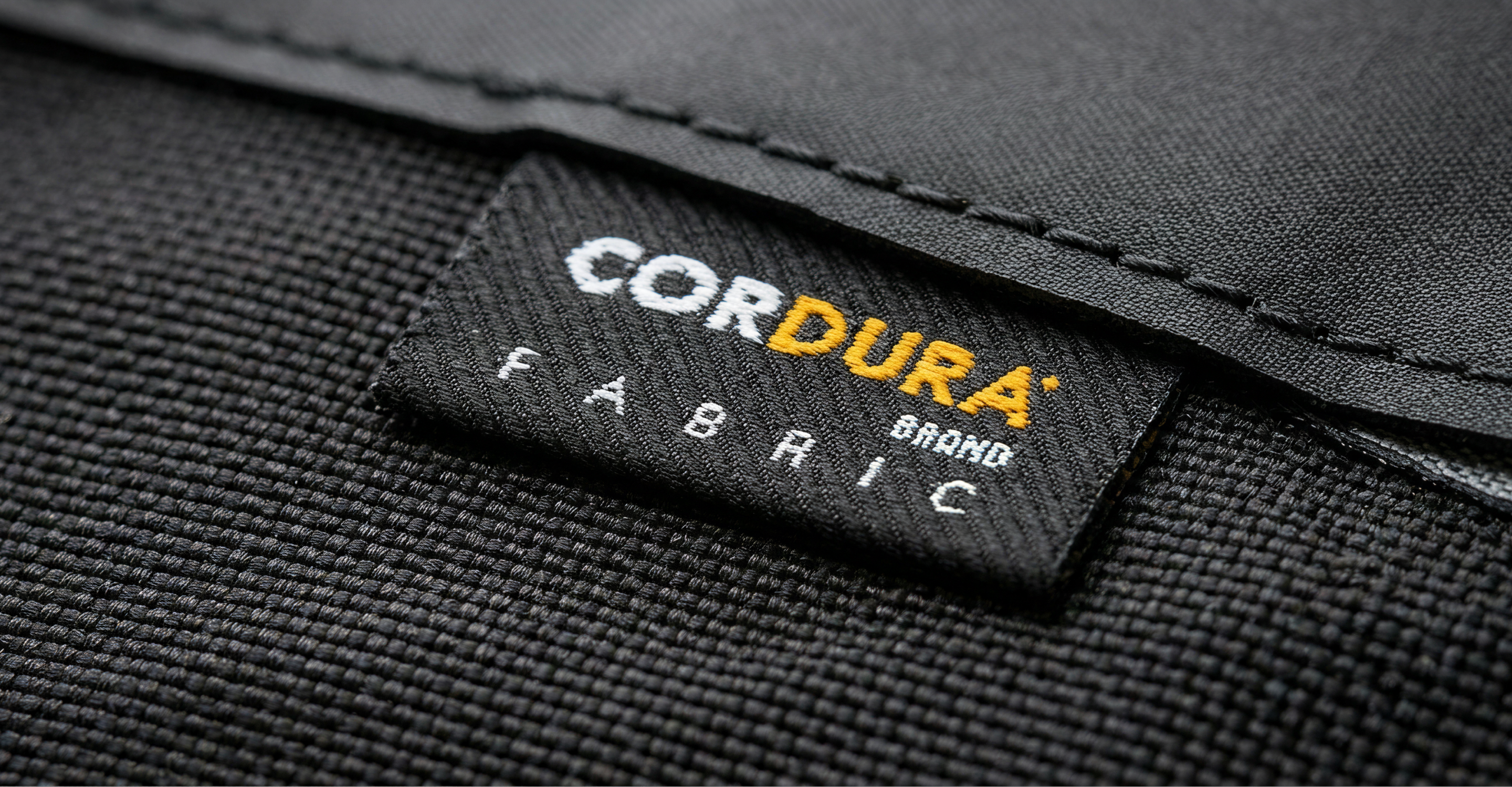 Close-up of a black fabric tag with white and yellow text reading CORDURA Brand Fabric, sewn onto a textured black material.