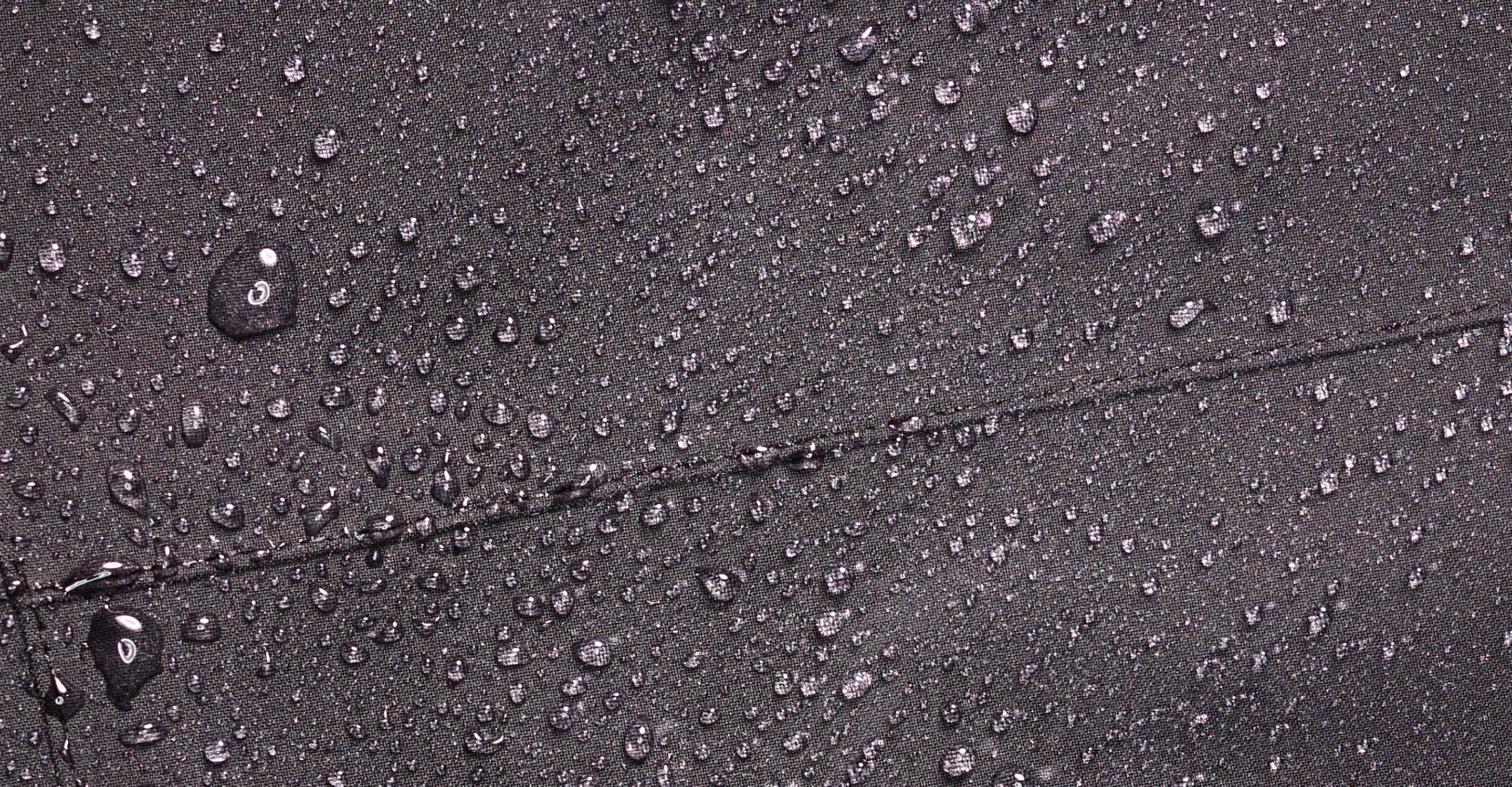 Close-up of water droplets on a dark, textured fabric surface, showing the materials water-resistant or waterproof properties.