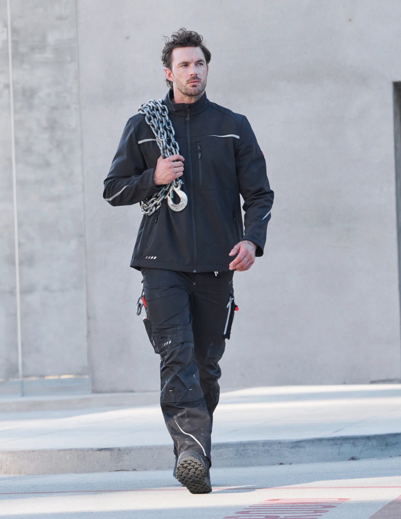 Man wearing a black work jacket and pants holding a chain, standing against a gray wall.