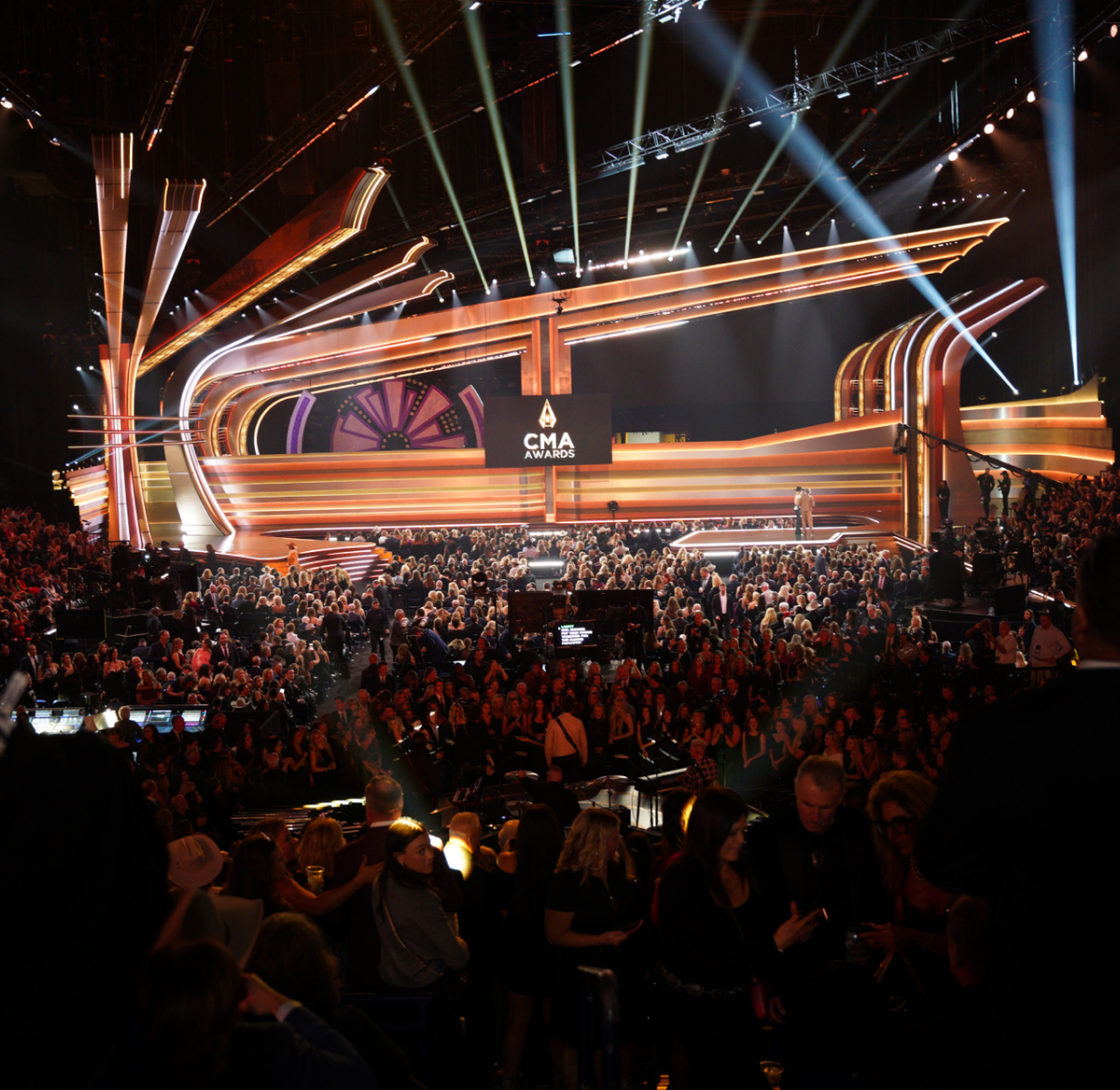A large crowd fills an arena facing an elaborate, brightly lit stage with CMA Awards displayed on a central screen. The atmosphere is lively, with people seated and awaiting the event.
