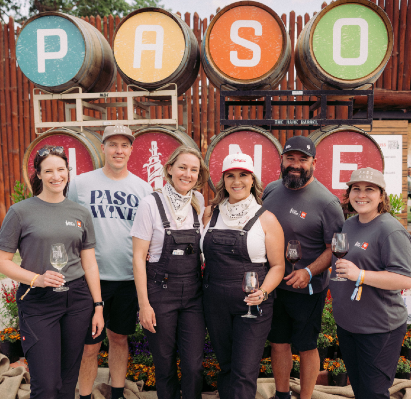 Six people pose smiling with wine glasses in front of large barrels spelling PASO WINE. They wear casual clothes and hats, with flowers and a wooden fence in the background.