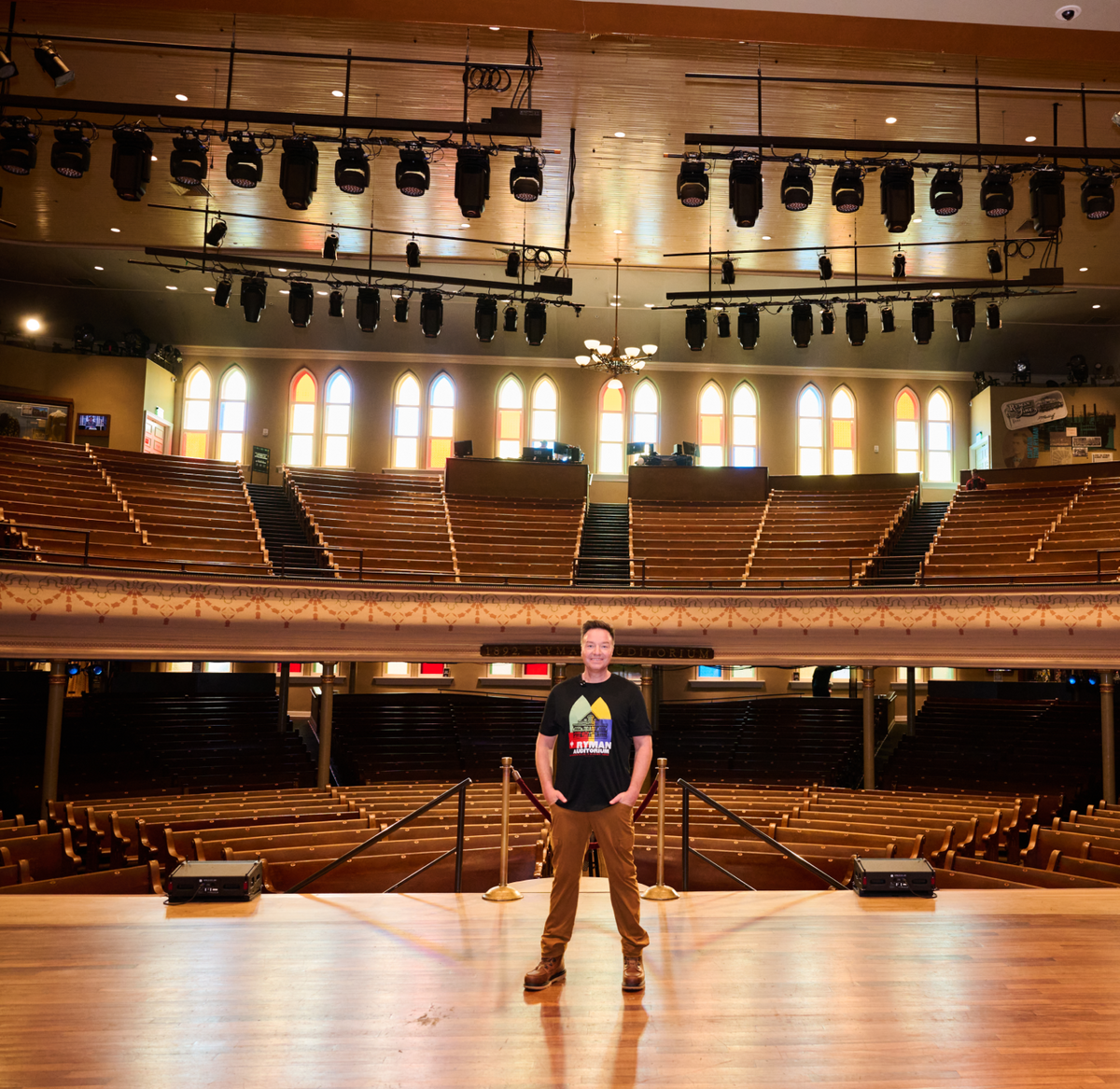 A person stands center stage in a large, empty theater with wooden seats and arched stained-glass windows. Stage lights hang above and rows of balcony seating are visible in the background.