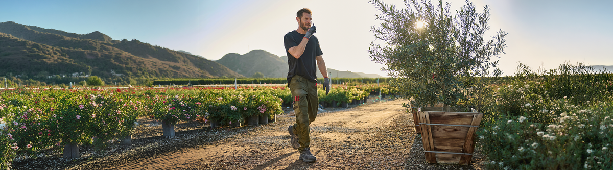 A man in casual clothes walks through a sunlit plant nursery with mountains in the background, holding a radio to his mouth. Trees and flowering bushes line the path.