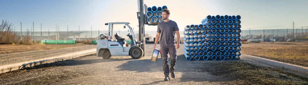 A man in work clothes carries a paint can and brush on a construction site, walking away from a forklift and large stacks of blue pipes under a clear sky.