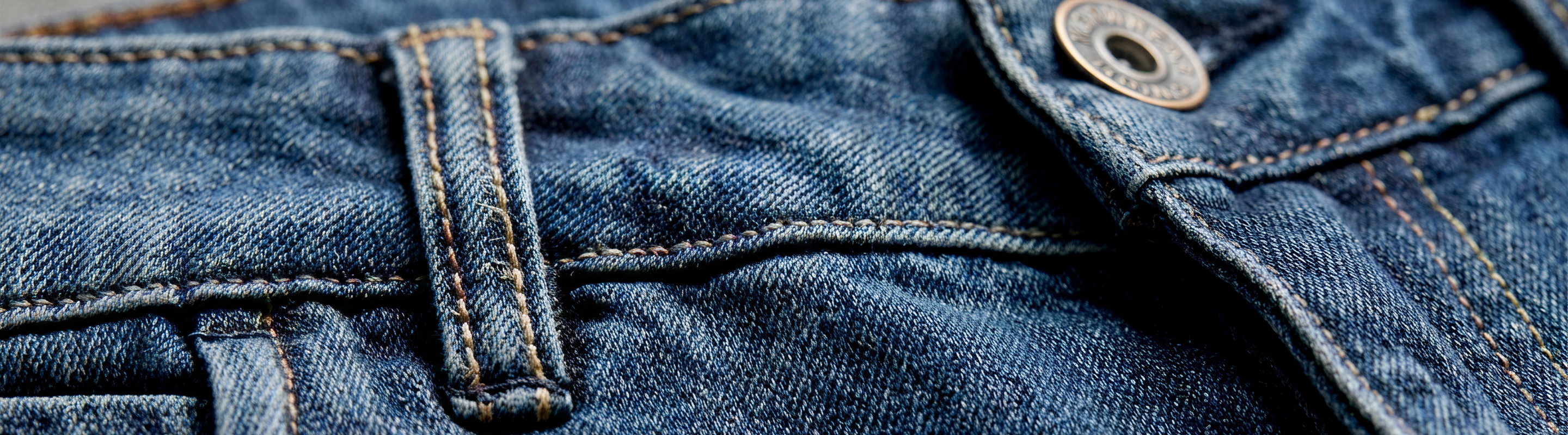 Close-up of blue denim jeans showing the waistband, belt loop, stitching, and a metal button.