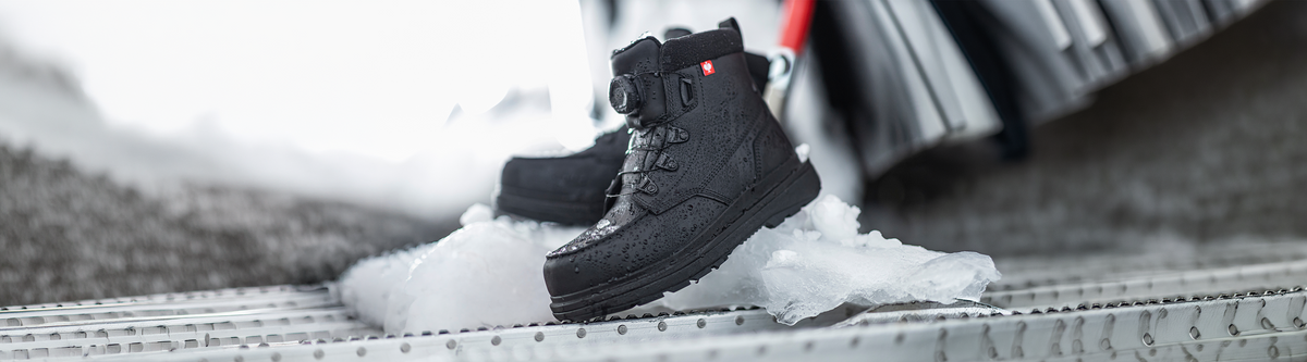 A pair of black winter boots with laces sits on ice-covered metal steps, with a blurred background suggesting a cold, outdoor environment.
