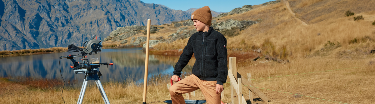 A person in a brown beanie and black jacket sits on a wooden fence near a lake, with mountains in the background and camera equipment set up on a tripod beside them in a grassy, outdoor area.