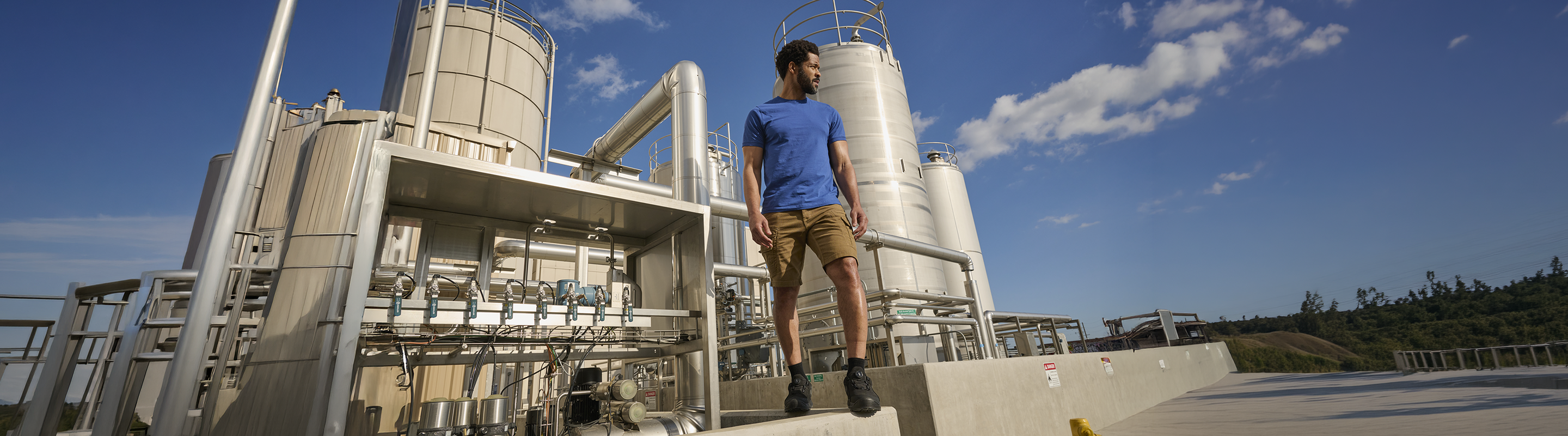 A man in a blue shirt and shorts walks on a concrete platform in front of large industrial tanks and pipes under a bright blue sky with some clouds.