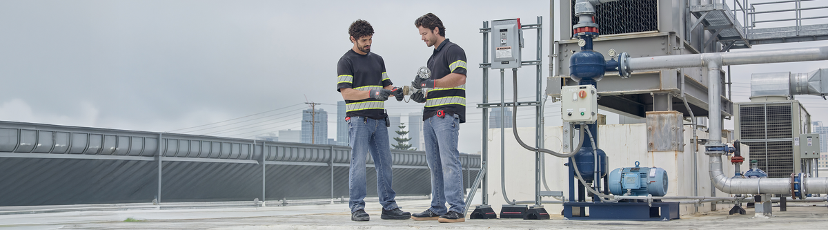 Two workers in safety gear inspect equipment on a rooftop industrial site, surrounded by pipes and machinery, with a city skyline visible in the background.