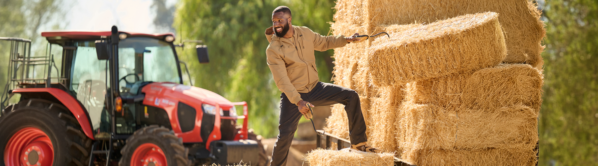 A man in a tan work jacket and dark pants stands on stacked hay bales next to a red tractor, holding a tool in an outdoor farm setting with trees in the background.