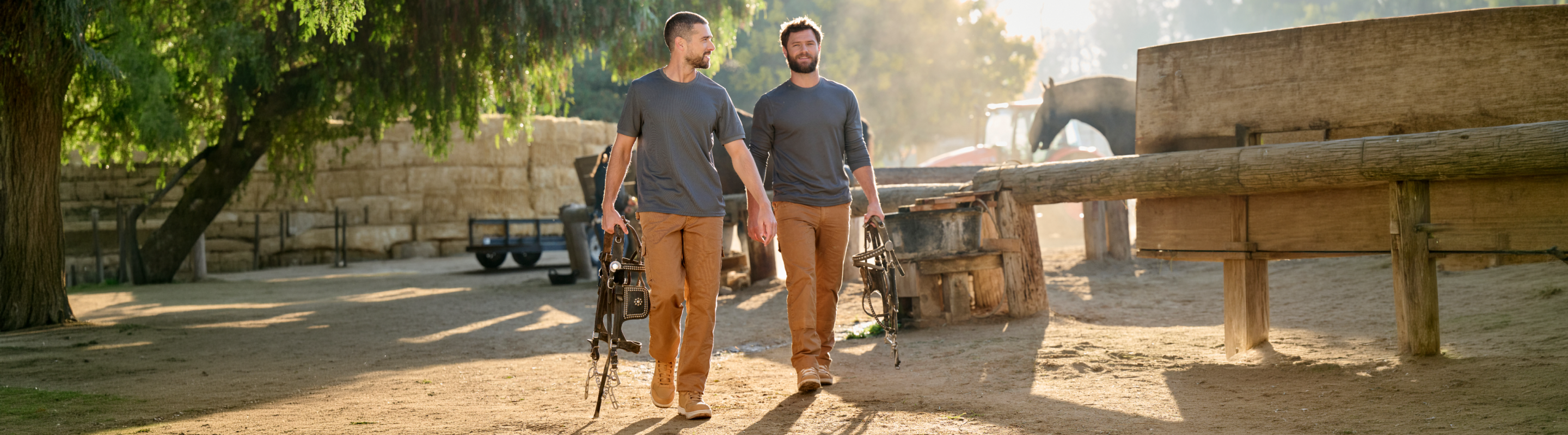 Two men in matching outfits walk outdoors on a sunny day, each holding a bow and arrows, with trees and rustic wooden structures in the background.