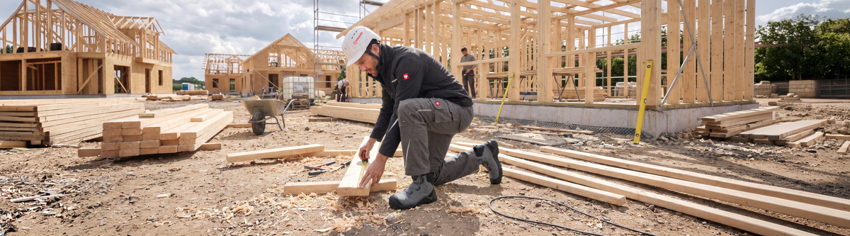 A man kneeling down to take measure of a wooden board. He is wearing a white hard hat, a black motion 2020 softshell jacket, grey cargo work pants motion 2020 and grey safety boots