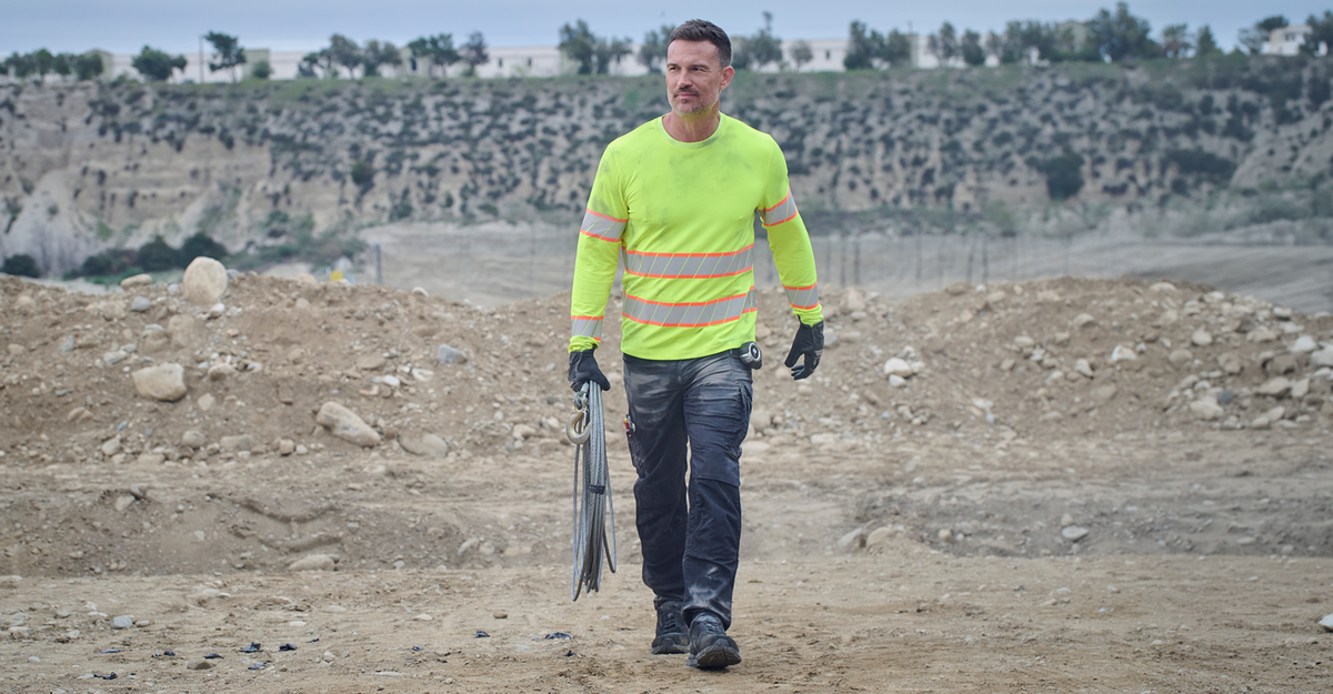 A man walking outdoors in a dusty construction site wearing a bright neon yellow high-visibility long-sleeve shirt with reflective silver stripes, dark gray work pants, and black gloves, while carrying a coiled cable.
