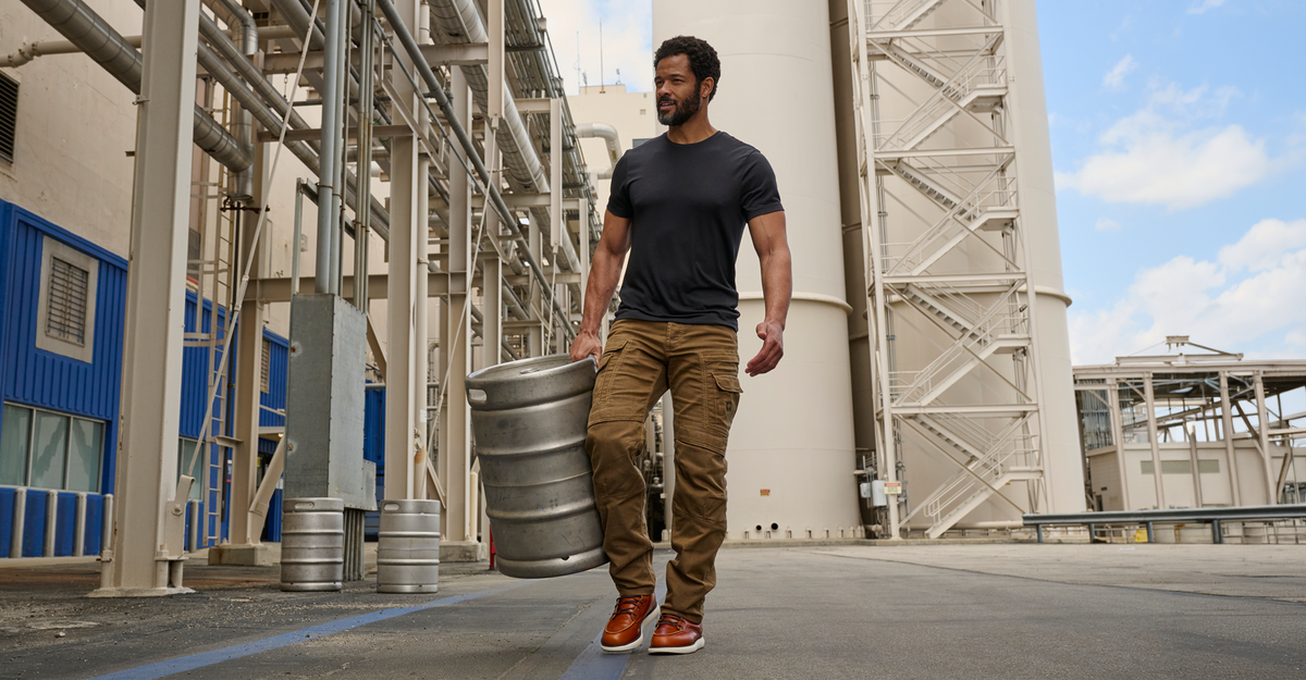 A man walking outdoors in an industrial area with large white tanks and metal structures, carrying a large silver keg barrel.