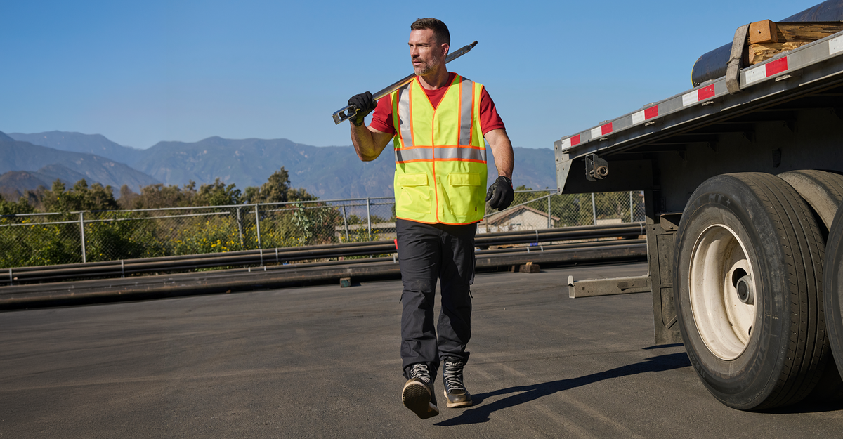 A male construction worker wearing a bright yellow safety vest over a red t-shirt walks on a highway shoulder next to a flatbed truck, carrying a long tool over his shoulder.