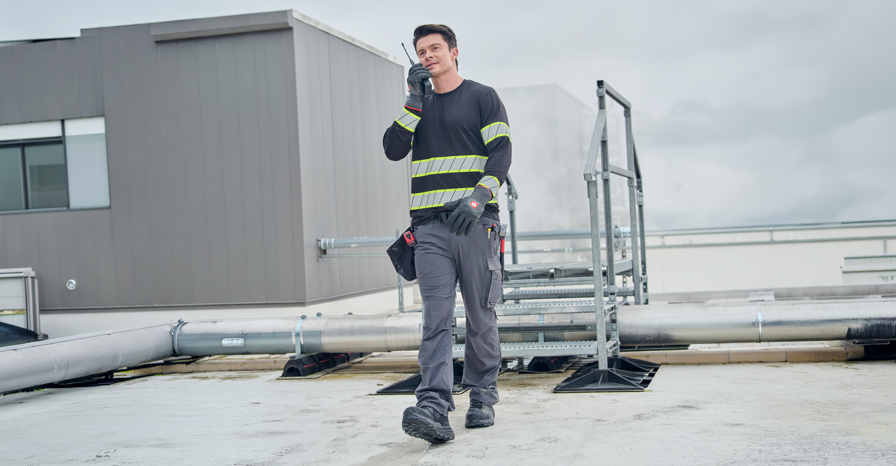 A male construction worker wearing a black high-visibility safety shirt with yellow reflective stripes, gray work pants, and black gloves walks on a rooftop while holding a radio walkie-talkie. A gray building and metal piping are visible in the background under an overcast sky.